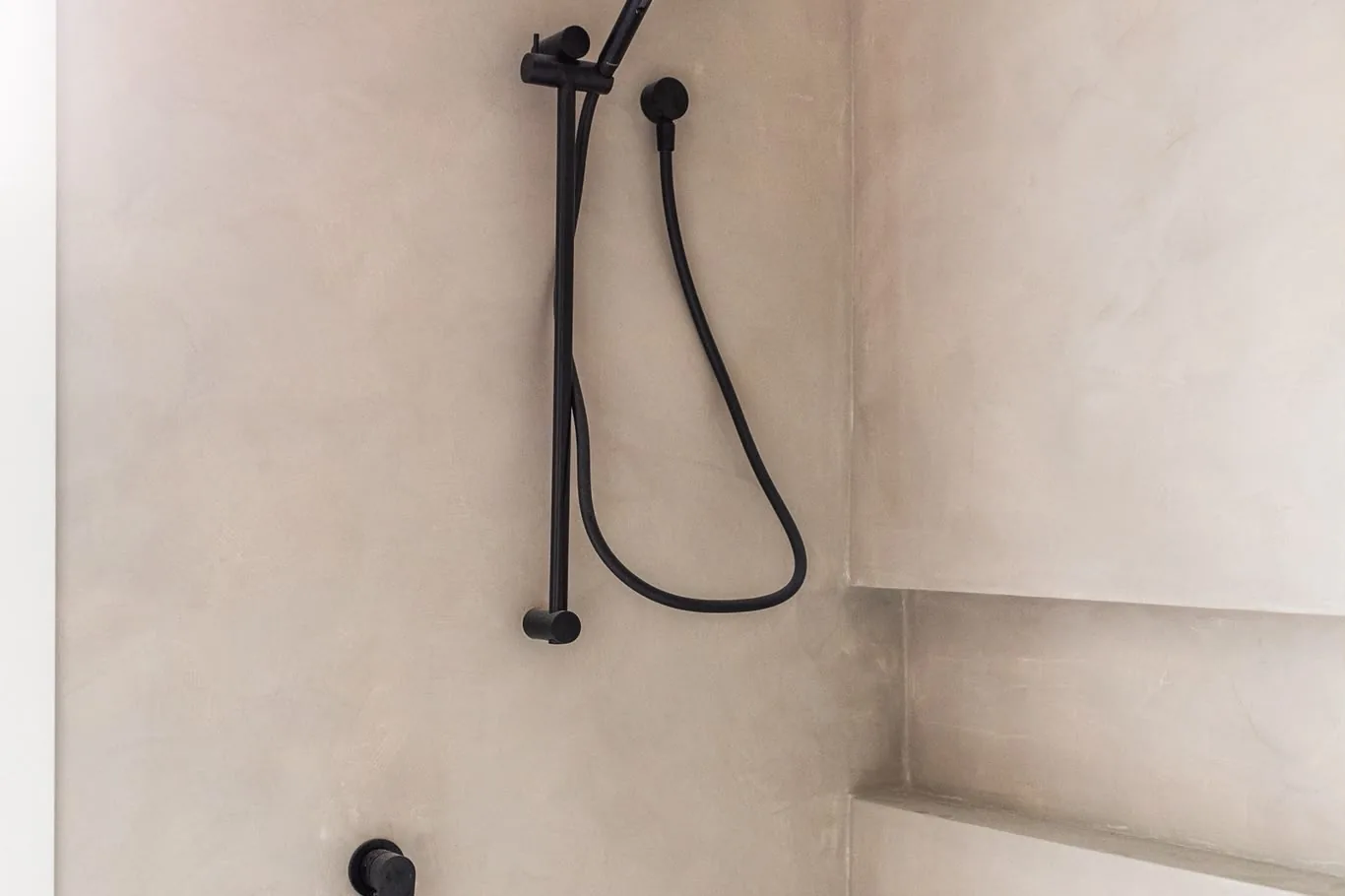 Pale microcement shower walls with a black hand shower on a rail and a long recessed shelf