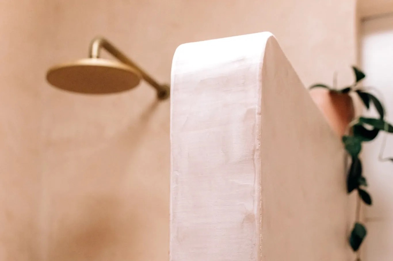 Shower in warm peach microcement with a brass shower head over a curved half-height wall
